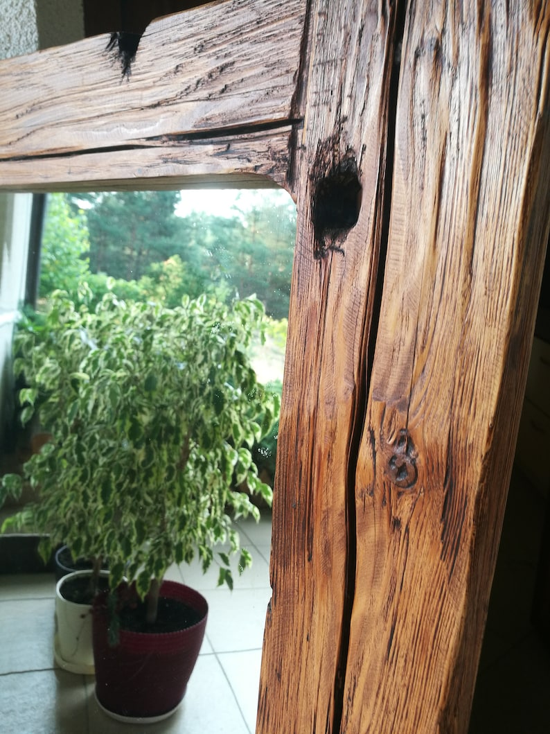 Puede incluir: Un marco de espejo de madera r&uacute;stico con un acabado de madera natural. El marco tiene un nudo y una grieta en la madera, lo que aumenta su car&aacute;cter. El espejo refleja una planta verde en una maceta.