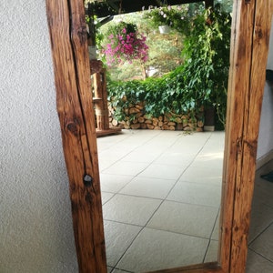 Puede incluir: Un espejo de madera r&uacute;stico con un marco rectangular. El marco est&aacute; hecho de madera gruesa y desgastada con una mancha marr&oacute;n oscura. El espejo refleja una vista de un patio con plantas en macetas y una pared de piedra.