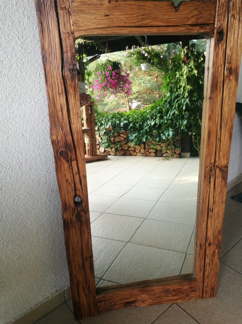 Puede incluir: Un espejo de madera r&uacute;stico con un marco rectangular. El marco est&aacute; hecho de madera gruesa y desgastada con una mancha marr&oacute;n oscura. El espejo refleja una vista de un patio con plantas en macetas y una pared de piedra.