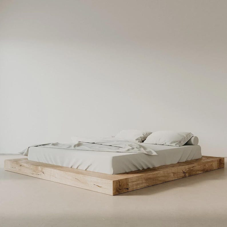 Puede incluir: Cama minimalista con colch&oacute;n y almohadas de color gris claro. La cama se asienta sobre una base de madera natural, creando una est&eacute;tica sencilla y moderna. La ropa de cama es de color neutro.