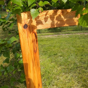 Op de afbeelding: Een rustieke houten wegwijzer met een natuurlijke houtnerf. De wegwijzer heeft een warme, goudbruine kleur en wordt gedeeltelijk bedekt door groene bladeren. Het staat tegen een achtergrond van groen gras en bomen.