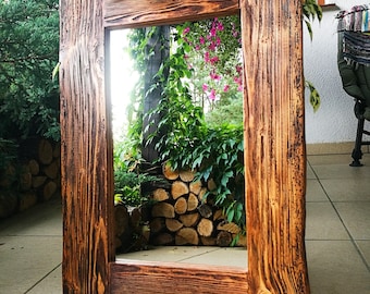 Op de afbeelding: Een rustieke houten spiegel met een rechthoekige lijst. De lijst is gemaakt van gerecycled hout met een donkerbruine afwerking. De spiegel reflecteert een uitzicht op groene planten en een stapel hout.