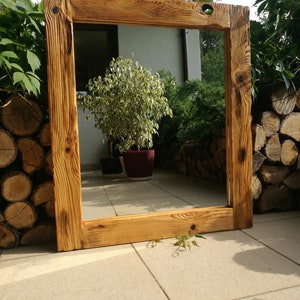 Op de afbeelding: Een grote, rechthoekige houten spiegel met een rustieke, natuurlijke houten lijst. De lijst heeft zichtbare houtnerf en knopen, met een warme, goudbruine kleur. De spiegel staat buiten, leunend tegen een muur.