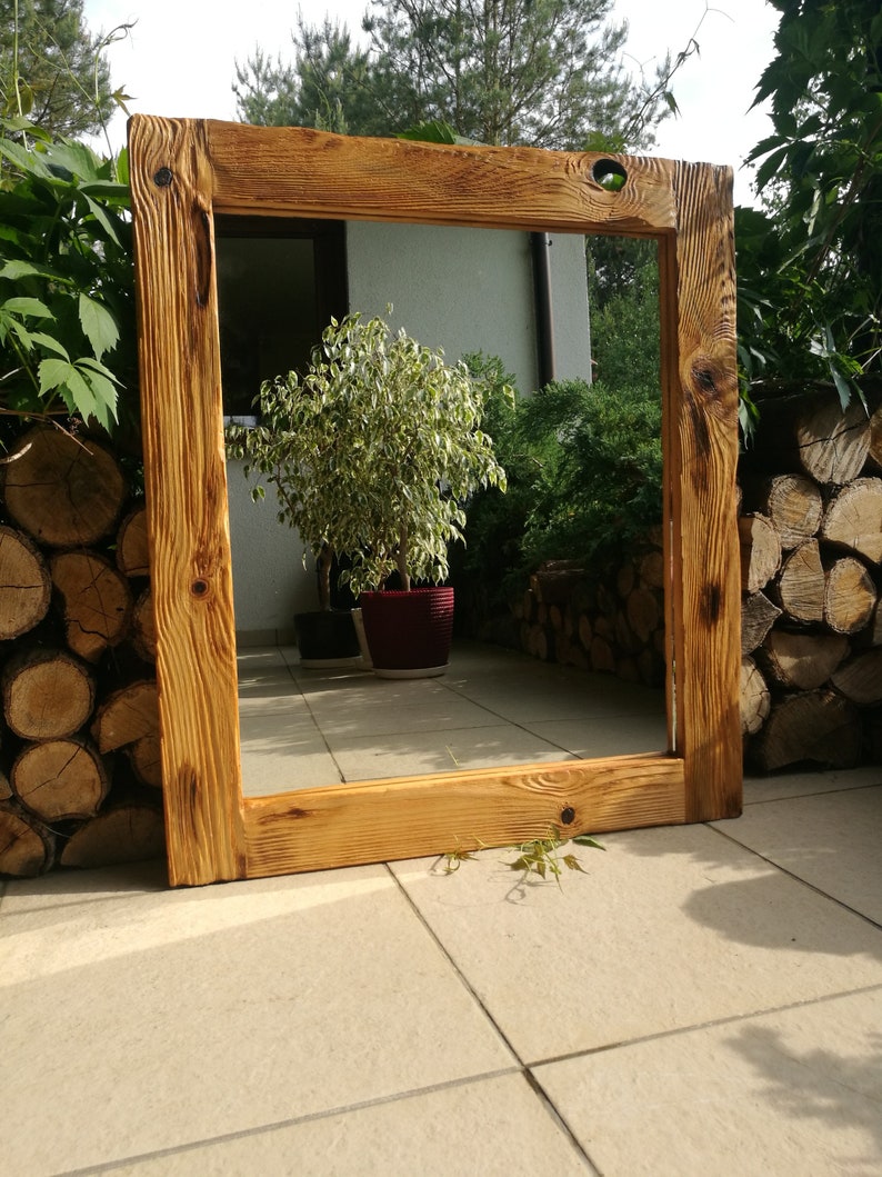 Op de afbeelding: Een grote, rechthoekige houten spiegel met een rustieke, natuurlijke houten lijst. De lijst heeft zichtbare houtnerf en knopen, met een warme, goudbruine kleur. De spiegel staat buiten, leunend tegen een muur.