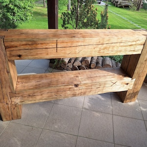 Puede incluir: Una estantería de madera rústica con dos estantes. La estantería está hecha de madera toscamente labrada con un acabado natural. La estantería está sobre un suelo de baldosas.