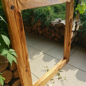 Op de afbeelding: Een rustiek, rechthoekig houten frame met een natuurlijke, lichtbruine afwerking. Het frame is gemaakt van dik, getextureerd hout, met zichtbare nerven en knopen. Het frame is leeg, wat suggereert dat het is ontworpen voor een spiegel of kunstwerk. Het frame bevindt zich buiten, met groen op de achtergrond.