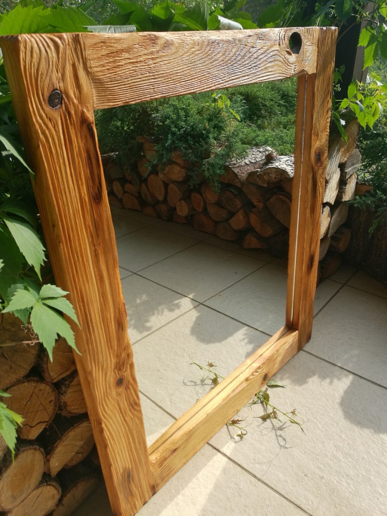 Op de afbeelding: Een rustiek, rechthoekig houten frame met een natuurlijke, lichtbruine afwerking. Het frame is gemaakt van dik, getextureerd hout, met zichtbare nerven en knopen. Het frame is leeg, wat suggereert dat het is ontworpen voor een spiegel of kunstwerk. Het frame bevindt zich buiten, met groen op de achtergrond.