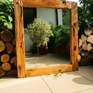 Op de afbeelding: Een grote, rechthoekige houten spiegel met een natuurlijke, rustieke afwerking. Het frame is gemaakt van dik, lichtbruin hout met zichtbare nerven en knopen. De spiegel wordt buiten tentoongesteld en weerspiegelt een tuin.