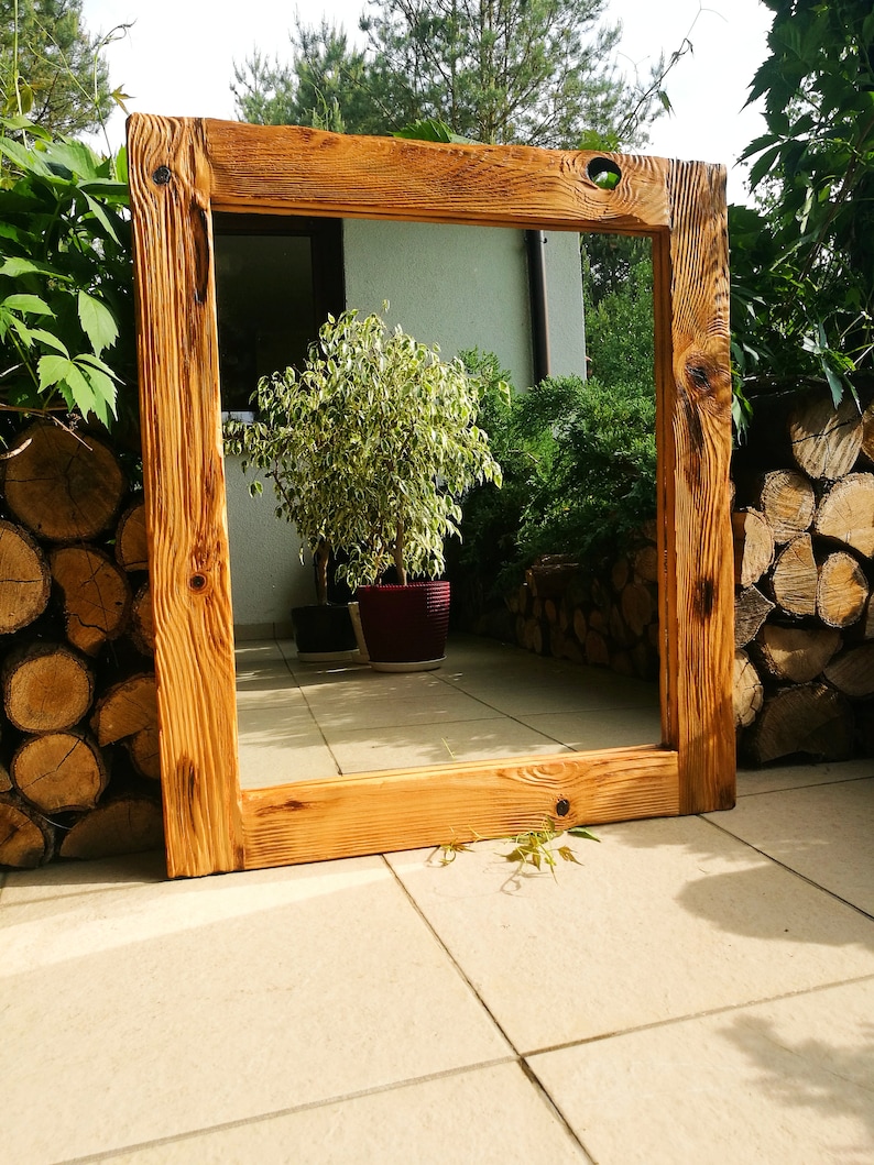 Op de afbeelding: Een grote, rechthoekige houten spiegel met een natuurlijke, rustieke afwerking. Het frame is gemaakt van dik, lichtbruin hout met zichtbare nerven en knopen. De spiegel wordt buiten tentoongesteld en weerspiegelt een tuin.