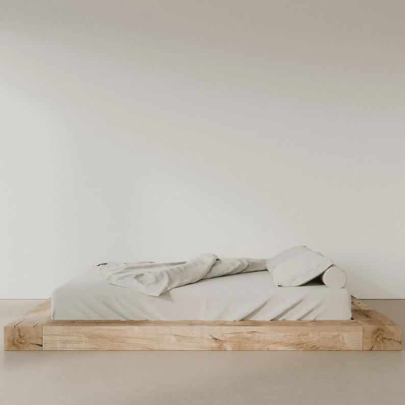 Puede incluir: Una cama de plataforma minimalista con colch&oacute;n y ropa de cama de color claro. La estructura de la cama es de madera natural en bruto. Se ve un coj&iacute;n cil&iacute;ndrico y una manta doblada sobre la cama. La habitaci&oacute;n tiene una paleta de colores neutros.