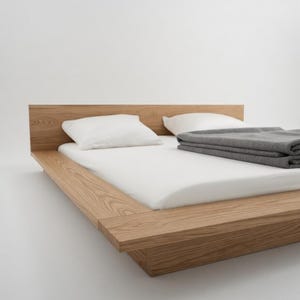 Puede incluir: Una cama de madera moderna con cabecero, colch&oacute;n blanco, dos almohadas blancas y una manta gris doblada. El marco de la cama es de madera clara y est&aacute; a baja altura, creando una est&eacute;tica minimalista.