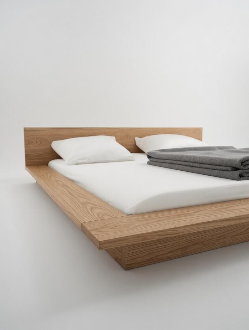 Puede incluir: Una cama de madera moderna con cabecero, colch&oacute;n blanco, dos almohadas blancas y una manta gris doblada. El marco de la cama es de madera clara y est&aacute; a baja altura, creando una est&eacute;tica minimalista.