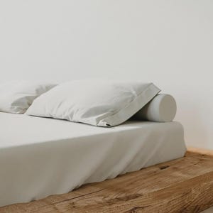 Puede incluir: Un juego de s&aacute;banas blancas que incluye una s&aacute;bana encimera, una funda de almohada rectangular y un coj&iacute;n cil&iacute;ndrico, todo sobre una base de cama de madera natural. La ropa de cama es de color blanco liso con una textura suave y mate.