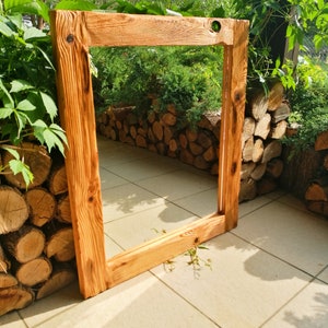 Op de afbeelding: Een groot, rechthoekig houten frame met een natuurlijke, lichtbruine afwerking. Het frame heeft zichtbare houtnerf en knopen, wat wijst op een rustieke uitstraling. Het frame leunt tegen een achtergrond van groen en opgestapelde boomstammen.