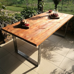 Op de afbeelding: Een houten tafel met een rustieke afwerking en metalen poten. De tafel is gedekt met een brood, fruit en een vaas met bloemen. De tafel staat op een betegeld terras.
