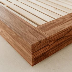Puede incluir: Primer plano de una estructura de cama de madera con acabado de madera natural. La estructura presenta un dise&ntilde;o rectangular robusto con vetas de madera visibles y una base de listones. La estructura de la cama es de madera maciza.