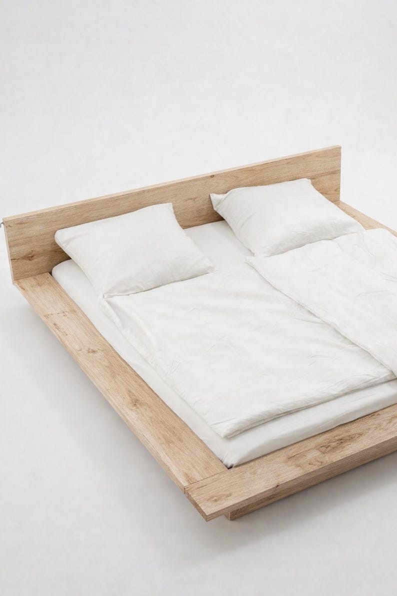 Puede incluir: Una cama moderna con estructura de madera clara y dise&ntilde;o minimalista. La cama est&aacute; hecha con ropa de cama blanca, que incluye una s&aacute;bana bajera, un edred&oacute;n y dos almohadas. La est&eacute;tica general es limpia y contempor&aacute;nea.