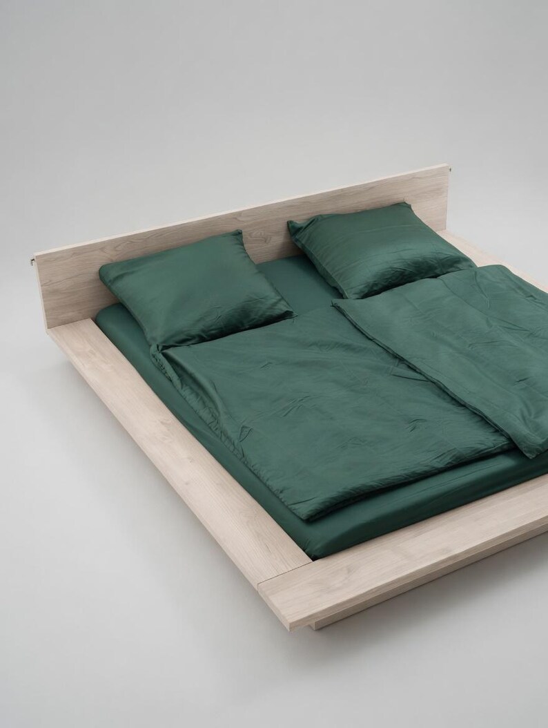 Puede incluir: Una cama con estructura de madera clara y ropa de cama verde oscuro. El juego incluye una s&aacute;bana bajera, dos almohadas y un edred&oacute;n. La cama est&aacute; sobre un fondo blanco liso.