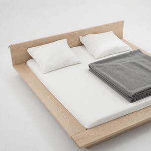 Puede incluir: Una cama de madera minimalista con un colch&oacute;n blanco, dos almohadas blancas y una manta gris doblada. La cama tiene un dise&ntilde;o simple y moderno, ideal para un dormitorio. El marco es de madera clara.