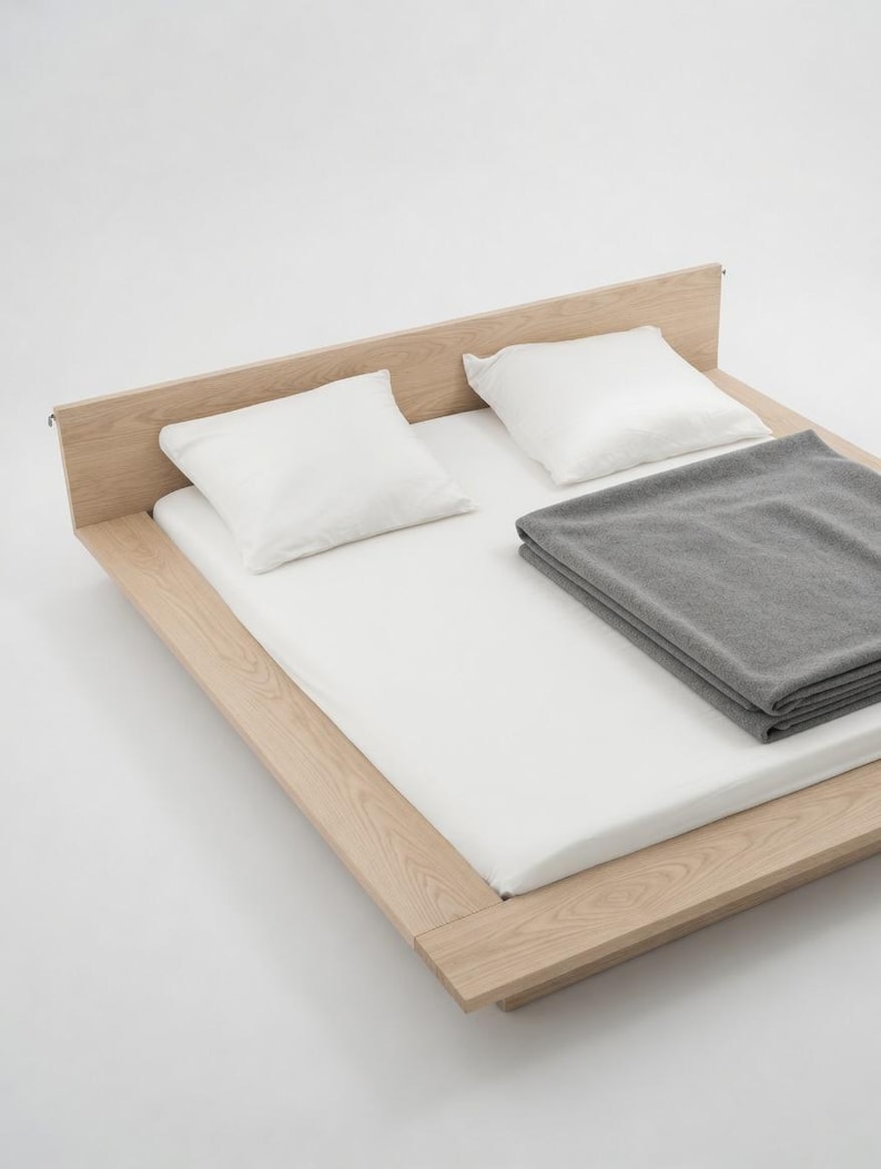 Puede incluir: Una cama de madera minimalista con un colch&oacute;n blanco, dos almohadas blancas y una manta gris doblada. La cama tiene un dise&ntilde;o simple y moderno, ideal para un dormitorio. El marco es de madera clara.