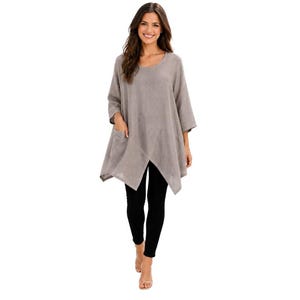 May include: A light taupe, loose-fitting tunic top with three-quarter length sleeves, a rounded neckline, and a distinctive asymmetrical crossover hemline featuring two front pockets, paired with solid black leggings.