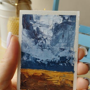 Puede incluir: Pequeña pintura de paisaje con pinceladas texturizadas. La obra presenta un cielo azul y blanco sobre un campo marrón dorado. Firmada en la esquina inferior izquierda. La pintura está sobre un pequeño lienzo rectangular.