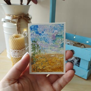 Puede incluir: Una pequeña pintura al óleo rectangular que representa un paisaje con una técnica de empaste texturizada. La obra de arte presenta un cielo azul y blanco, un campo amarillo y marrón, y vegetación verde. Las iniciales del artista, "N.S.", son visibles en la esquina inferior izquierda.