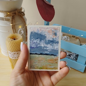 Könnte beinhalten: Miniatur-Landschaftsbild im impressionistischen Stil. Das Kunstwerk zeigt einen Himmel in Blau und Weiß über einem Feld in Grün und Orange. Die Initialen des Künstlers, "N.S.", sind in der unteren linken Ecke zu sehen.
