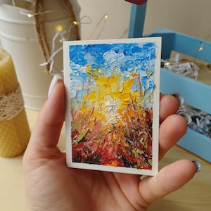Puede incluir: Pequeña pintura al óleo rectangular con una textura impasto. La obra de arte representa un paisaje con un cielo azul y blanco brillante sobre un campo de amarillo, naranja y rojo. La pintura se sostiene en una mano.