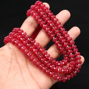 May include: A multi-strand necklace composed of small, round, translucent red beads. The beads are strung together with white string, creating a striking contrast. The necklace is displayed against a dark background.