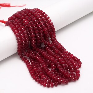 May include: Strands of deep red beads, likely gemstones, are displayed against a white background. The beads are small, round, and strung together, creating a cascading effect. The color is a rich, saturated red, and the beads have a slightly glossy finish.