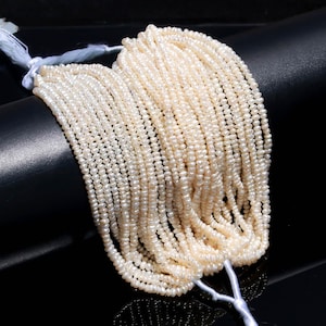 Può includere: Una collezione di piccole perle rotonde, di colore bianco crema, infilate insieme. I fili sono legati con un filo bianco, creando un fascio. Le perle hanno una lucentezza naturale e sono esposte su uno sfondo scuro.