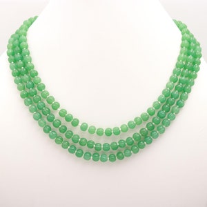 Peut inclure: Un collier à plusieurs rangs avec des rangées de perles vert clair. Les perles sont espacées uniformément et enfilées sur un cordon blanc. Le collier est présenté sur un mannequin blanc, mettant en valeur son design superposé.