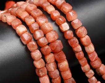 May include: A strand of orange sunstone beads, each bead is faceted and has a square shape. The beads are strung on a cord and are arranged in a row.