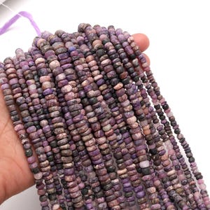 Puede incluir: Hebras de cuentas de piedras preciosas moradas y grises. Las cuentas son pequeñas, cilíndricas y tienen una forma ligeramente irregular. El color varía del lavanda claro al ciruela oscuro, con algunos tonos grises y marrones. Las cuentas están ensartadas en un hilo morado.