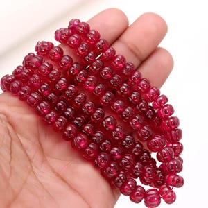 May include: Strands of deep red, carved gemstone beads. The beads have a rounded shape with intricate floral-like carvings. The beads are held in a hand, showcasing their color and detail. The gemstones appear to be polished and translucent.