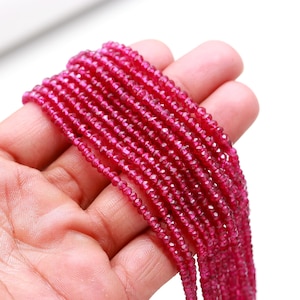 May include: Strands of small, faceted, deep red beads. The beads are held in a hand, showcasing their color and texture. The beads appear to be strung together, suggesting they are for jewelry making or crafting.