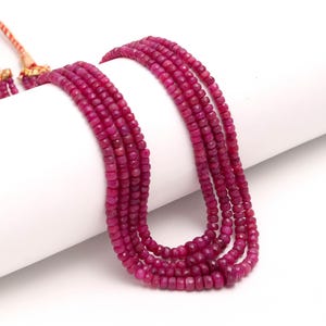 Op de afbeelding: Een meerstrengs ketting met kleine, gefacetteerde, dieproze robijnkralen. De ketting is over een wit oppervlak gedrapeerd, waardoor de levendige kleur en textuur van de edelstenen worden benadrukt. De ketting heeft een goudkleurige sluiting.