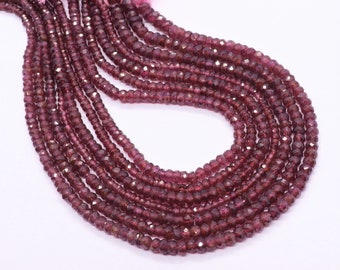 May include: Strands of faceted, deep red garnet beads. The beads are small and uniform in size, with a polished, reflective surface. The strands are arranged in a flowing, curved pattern against a white background. The beads are strung together, ready for jewelry making.