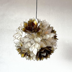 Puede incluir: Una lámpara colgante esférica con un diseño en capas. La luz presenta una mezcla de elementos en forma de pétalos blancos, dorados y marrones. La lámpara cuelga de un cable negro sobre un fondo blanco.