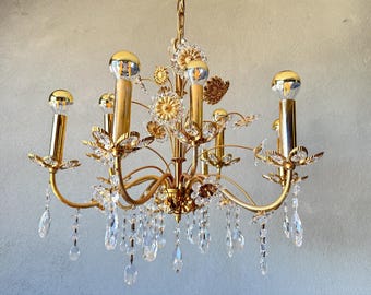 May include: A gold-toned chandelier with six light fixtures. The chandelier features floral and leaf accents, clear crystal droplets, and gold-colored bulb covers. The light fixtures are arranged in a circular pattern, creating a decorative lighting fixture.