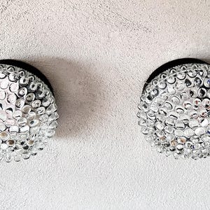 Op de afbeelding: Twee ronde, heldere glazen lampen met een bubbelachtige textuur. Elke lamp is gemonteerd op een zwarte basis en staat tegen een getextureerde, gebroken witte muur. Binnenverlichting.