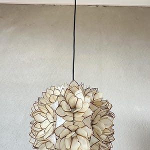 Op de afbeelding: Hanglamp met een bloemmotief. De lamp heeft een zwart snoer en plafondbevestiging. De lampenkap bestaat uit talrijke crèmekleurige, bloemblad-vormige elementen met bruine randen, die een bol vormen.