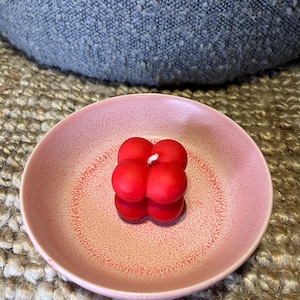 May include: A red bubble candle with a white wick sits on a pink speckled ceramic dish. The candle is made of four spheres fused together. The dish is round and has a textured surface.