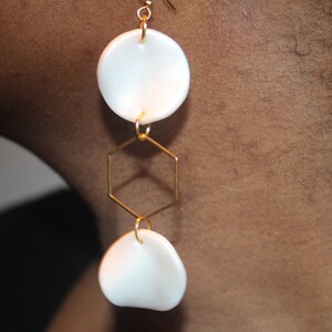 May include: White ceramic earrings with a gold hexagon charm. The earrings are dangling and have a simple, modern design.
