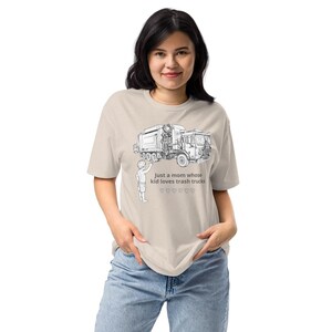 Pode incluir: Camiseta bege com uma ilustração em preto e branco de um caminhão de lixo e uma criança. O texto diz: "Just a mom whose kid loves trash trucks" com símbolos de coração. A camiseta é usada por uma pessoa.