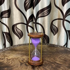Puede incluir: Un reloj de arena de madera con arena morada. El reloj de arena tiene un centro de cristal transparente y soportes de metal color oro. La parte superior e inferior de madera son de color marrón oscuro. El reloj de arena está sobre una mesa de madera.