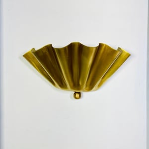 May include: A semi-circular wall sconce featuring a metallic gold finish with a pleated, fan-like design. The fixture has a wide, scalloped top edge and a small, spherical decorative element centered at the bottom point of the structure.
