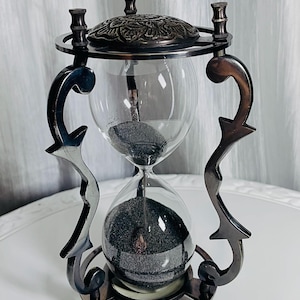 May include: An hourglass with a dark metallic frame featuring ornate, curved side supports and a circular base and top. The top piece has a decorative embossed pattern. The glass bulbs contain fine black sand, with some sand currently flowing.