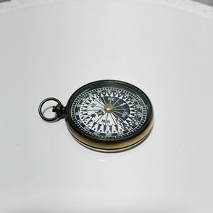 Puede incluir: Una brújula de bolsillo redonda con carcasa de metal color latón y esfera negra. El dial presenta marcas blancas, incluyendo los puntos cardinales N, S, E, O y grados numéricos del 0 al 360. Tiene un pequeño anillo de metal incorporado.